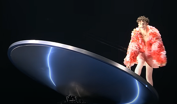 Nemo Nemo, in pink skirt and feathery jacket, climbing up a round whirligig see-saw thing.